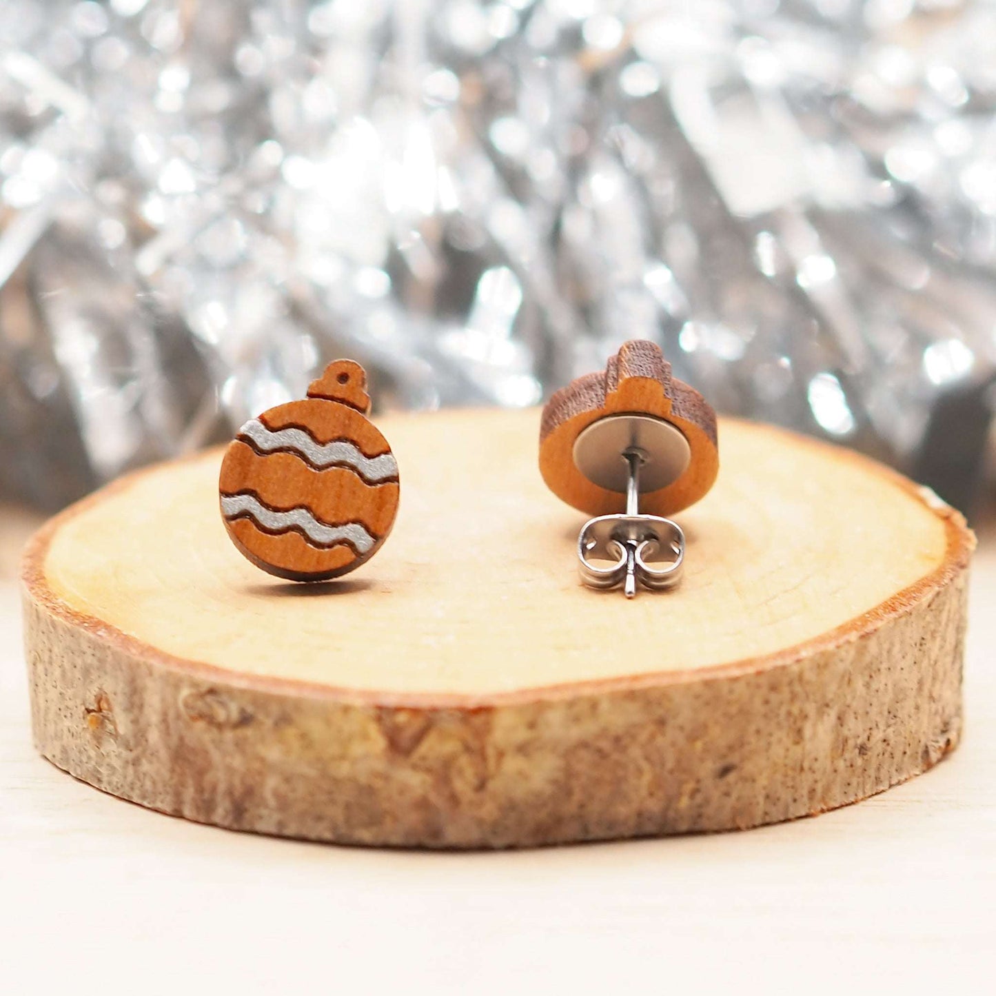 Silver Christmas bauble earrings with hand-painted silver pattern on wooden studs, displayed on a log slice.