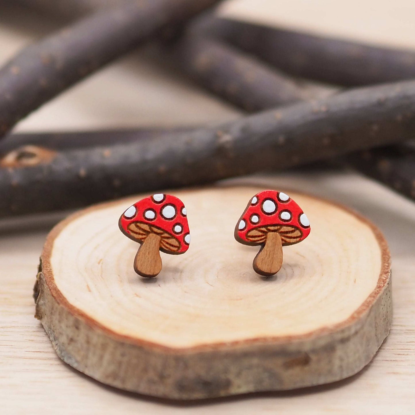 Red and white hand-painted mushroom stud earrings made from cherry wood with surgical steel backs, displayed on a wooden surface.
