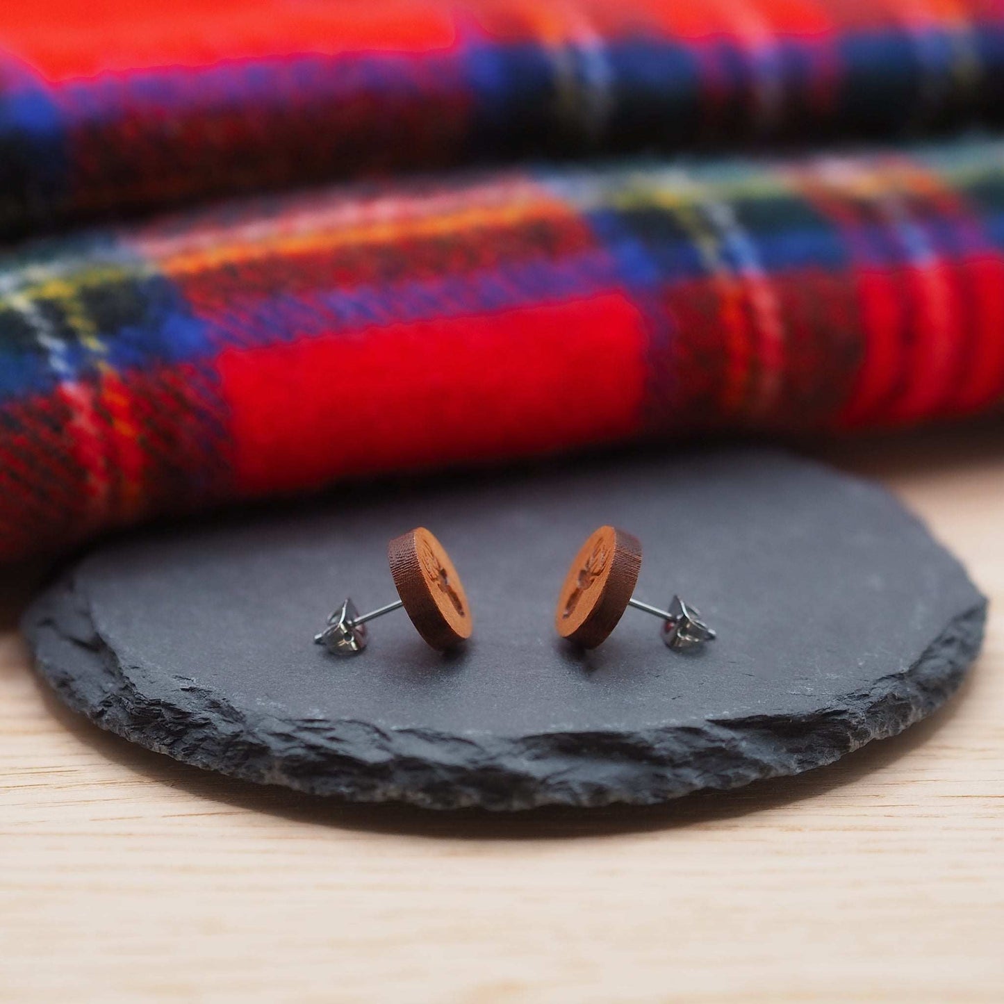 Stag head stud earrings made from cherry wood with surgical steel posts, displayed on a slate with a tartan fabric background.