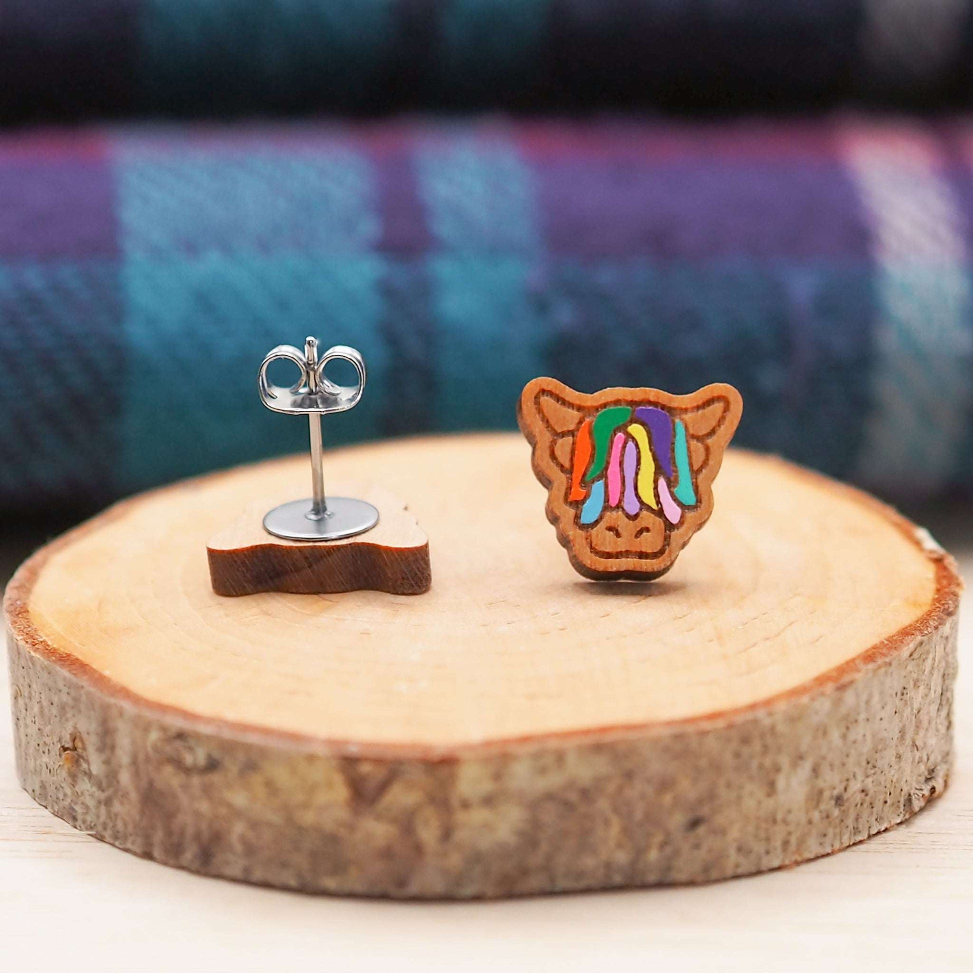 Multi-coloured Highland Cow stud earrings made from cherry wood and surgical steel on wooden display.