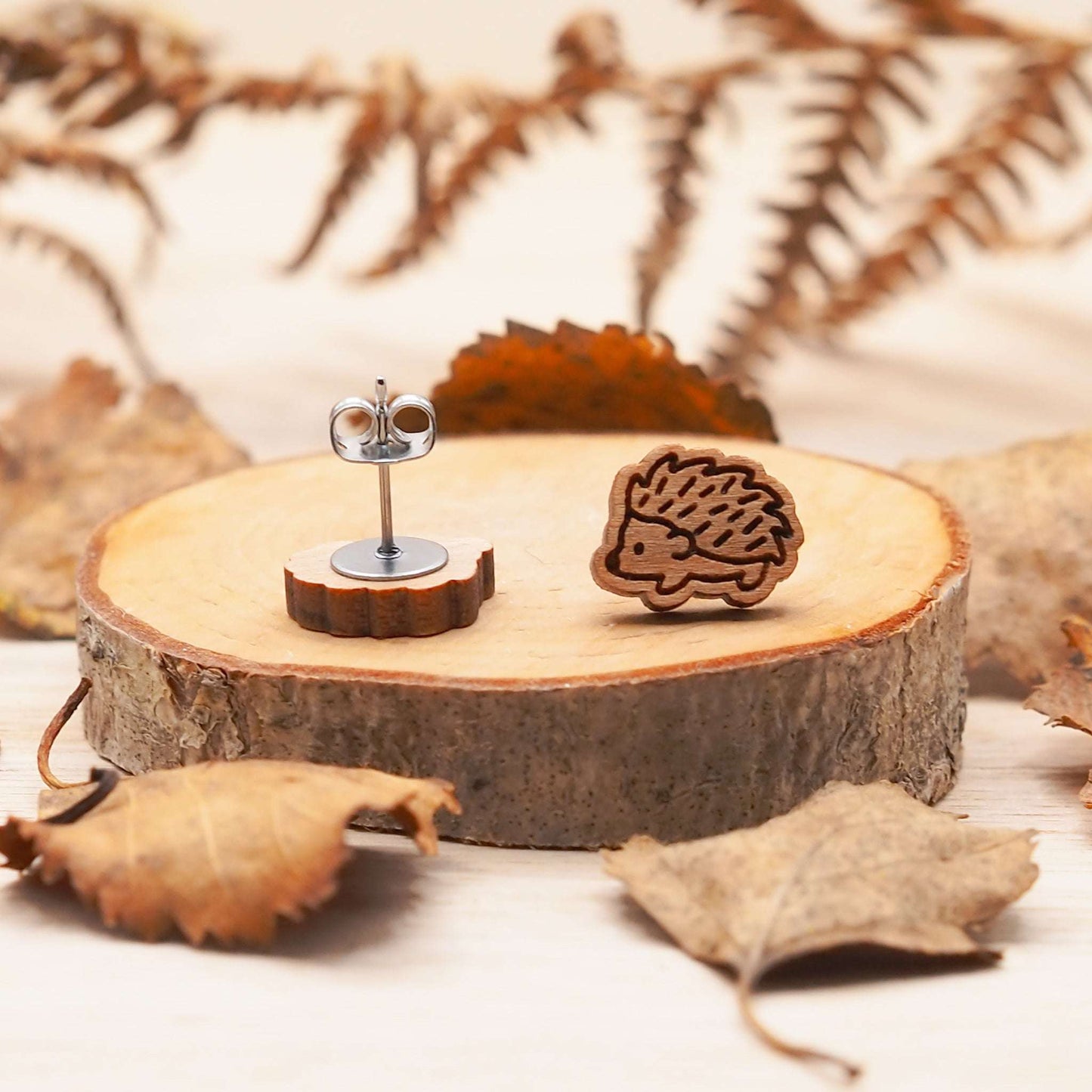 Wooden hedgehog stud earrings on a slice of wood surrounded by leaves, engraved on cherry wood with surgical steel backs.