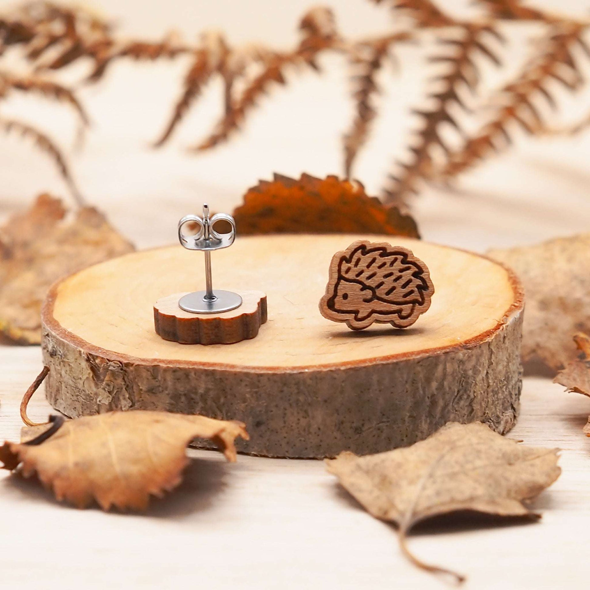 Wooden hedgehog stud earrings on a slice of wood surrounded by leaves, engraved on cherry wood with surgical steel backs.