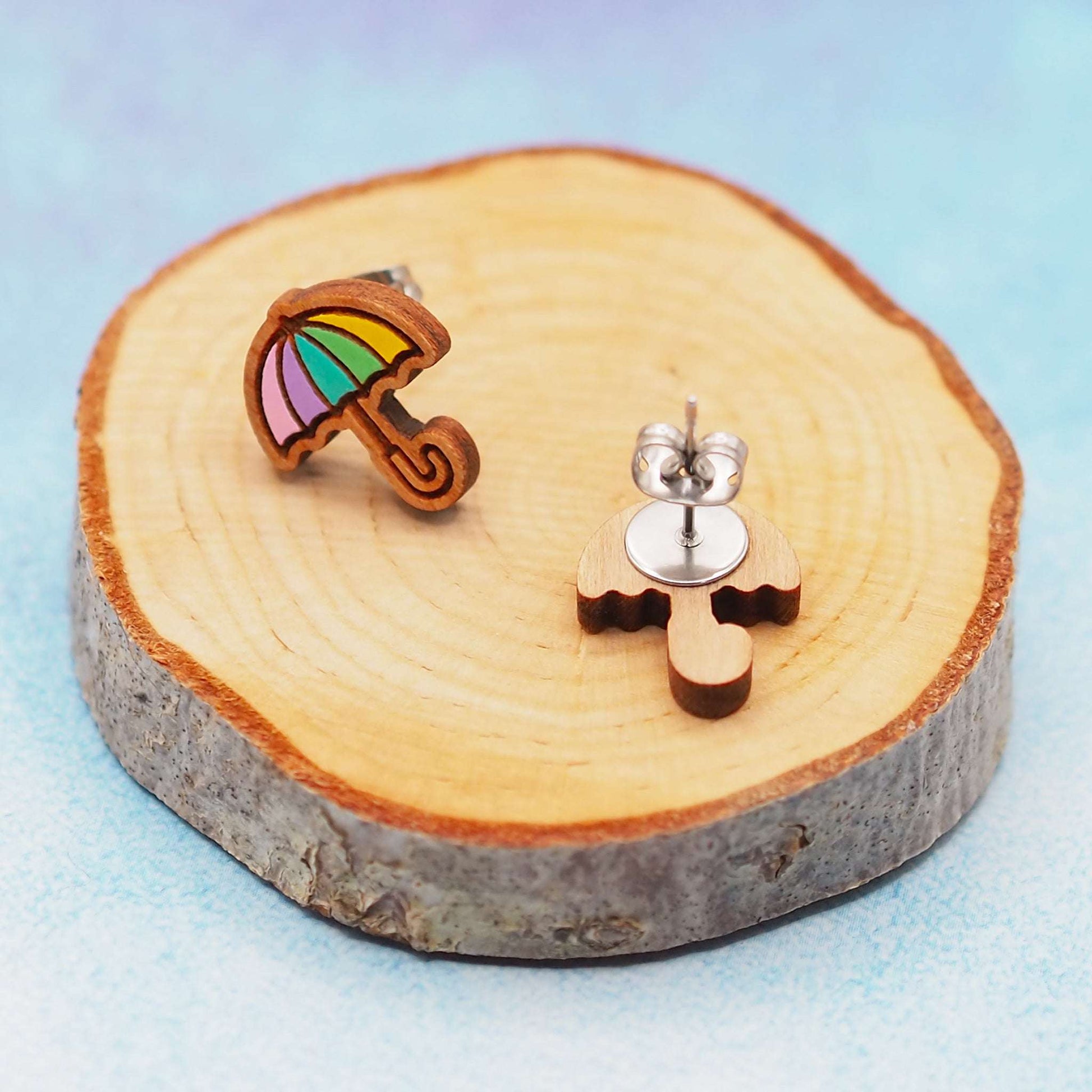 Pastel rainbow umbrella wooden stud earrings on decorative wood slice with steel backs.