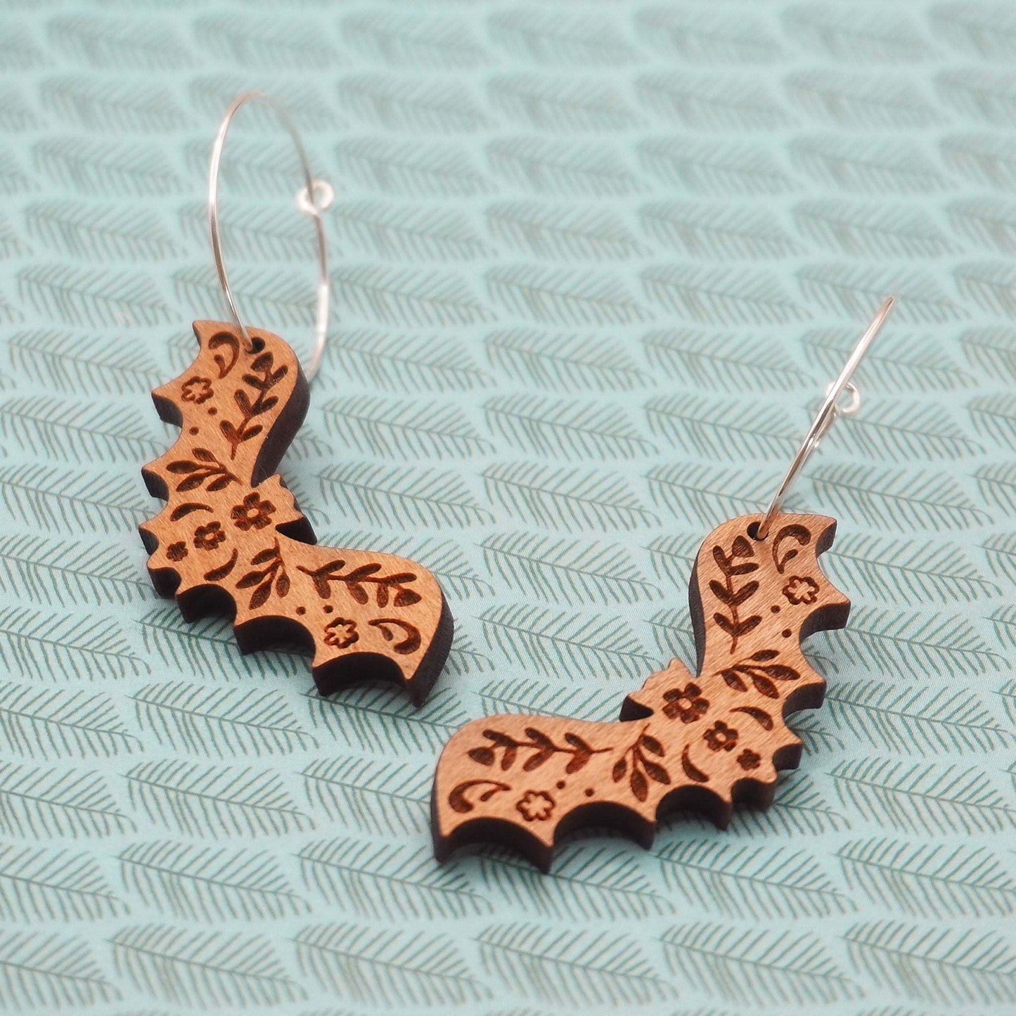 Laser cut bat hoop earrings made from cherry wood with sterling silver hoops on a patterned background.