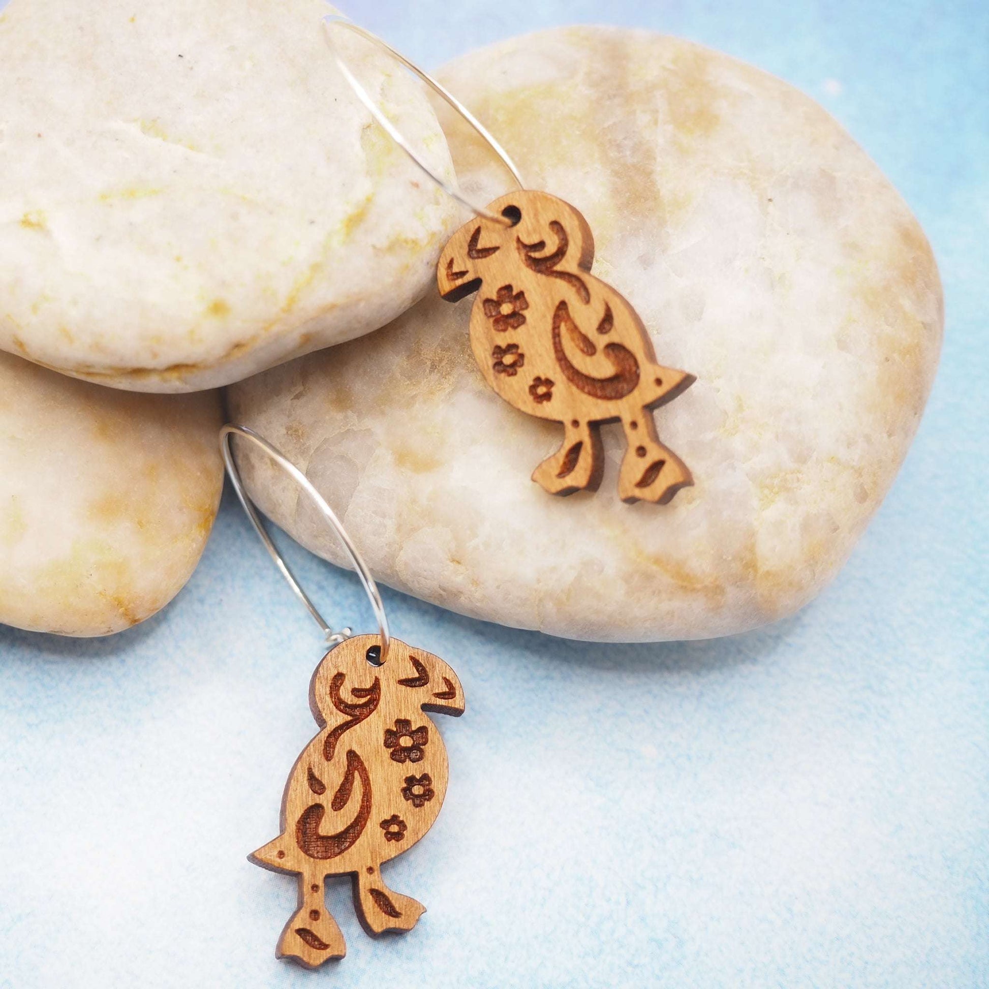 Wooden puffin engraved hoop earrings on sterling silver hoops, featuring a folk art design, displayed on smooth stones.