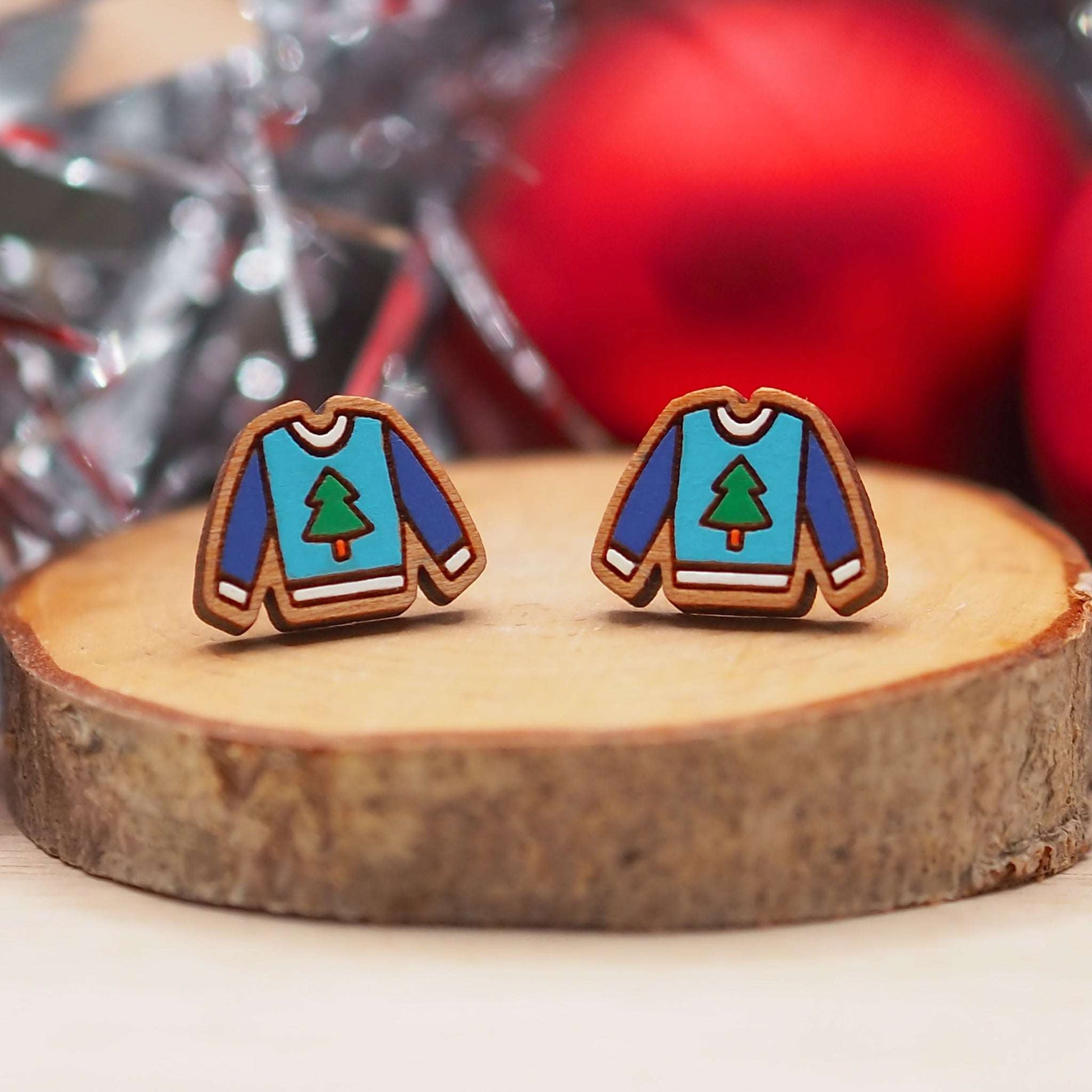 Christmas jumper stud earrings made from cherry wood and mounted on surgical steel posts, displayed on a wood slice with festive decor in the background.