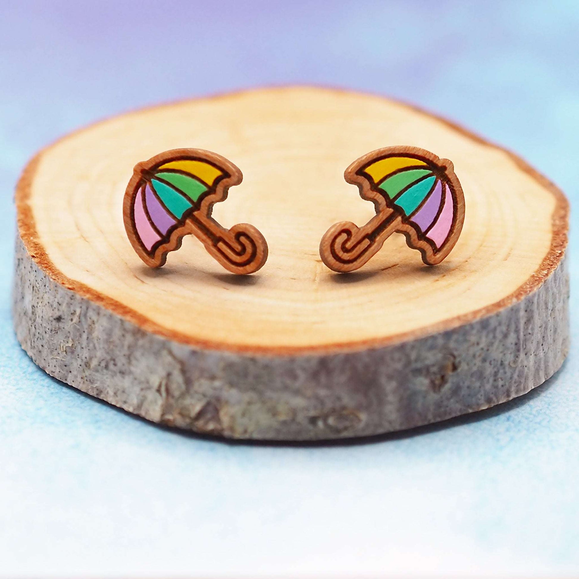 Pastel rainbow umbrella wooden stud earrings on a wooden slab with a blue background.