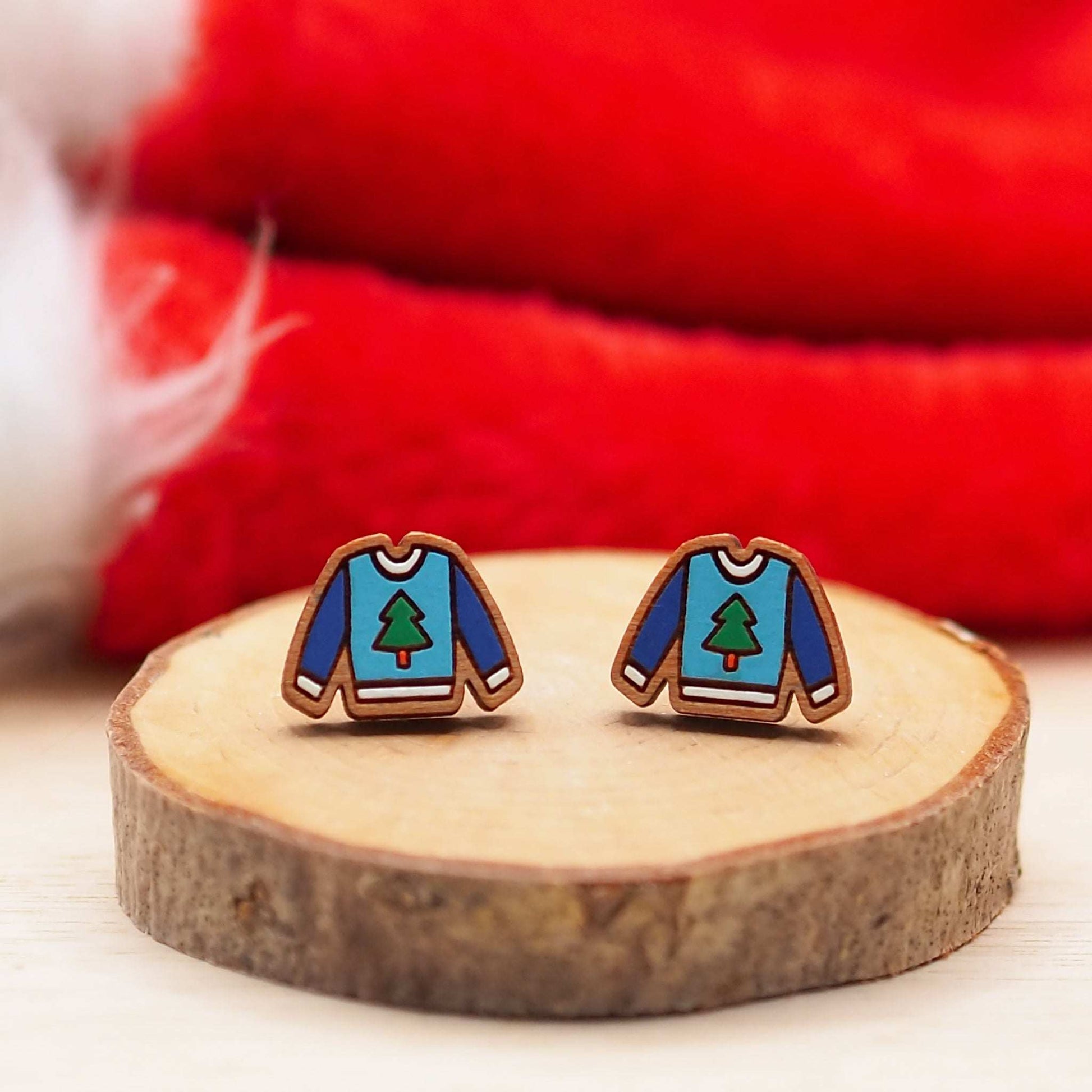 Cherry wood Christmas jumper stud earrings with tree design, mounted on surgical steel posts, displayed on a wooden surface with festive red background.