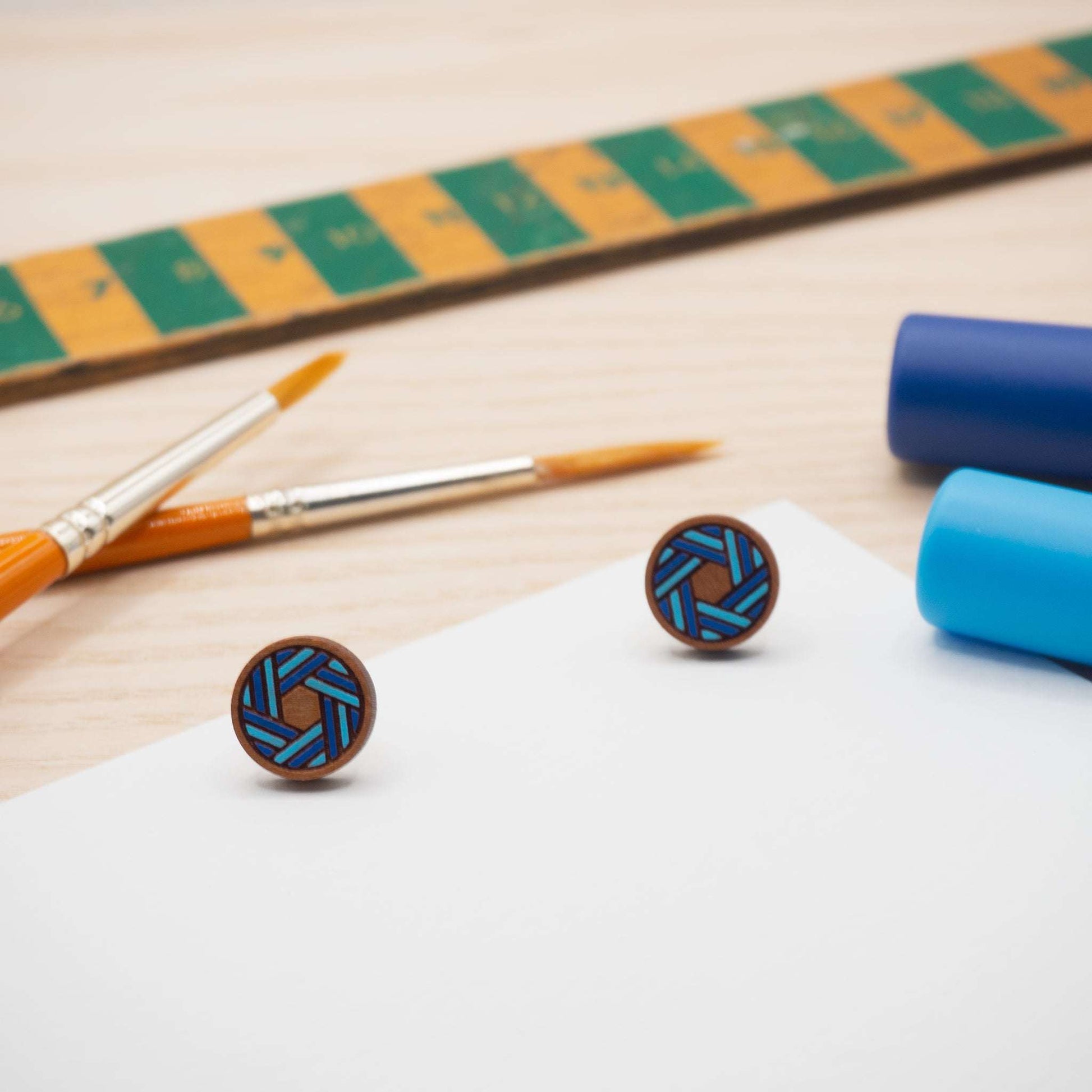Blue knot painted stud earrings on wooden background with paintbrushes and blue paint.