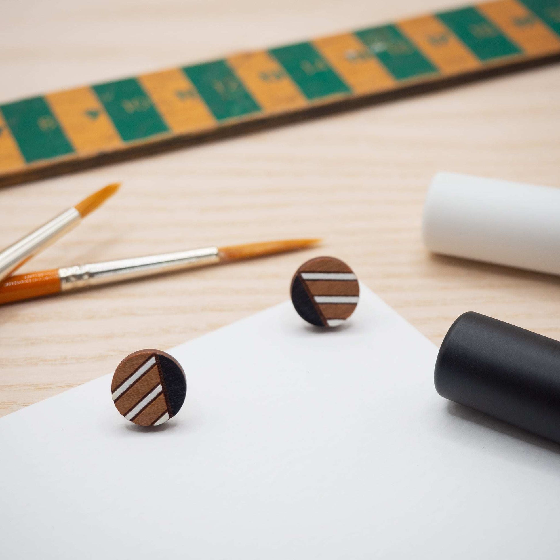 Monochrome geometric painted stud earrings made from cherry wood with black and white stripes, displayed on a wooden background with paintbrushes.