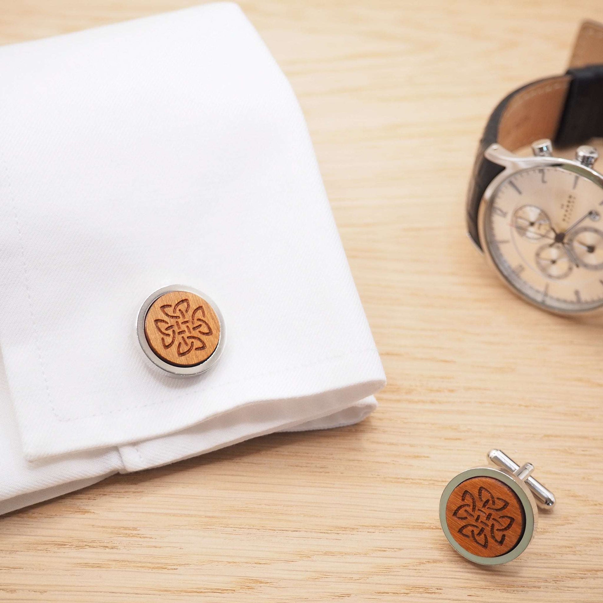 Celtic knot wooden cufflinks on white cuff with watch on wooden surface.